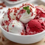 Scoop Of Vibrant Red Velvet Cake Ice Cream With Visible Cake Chunks In A White Bowl